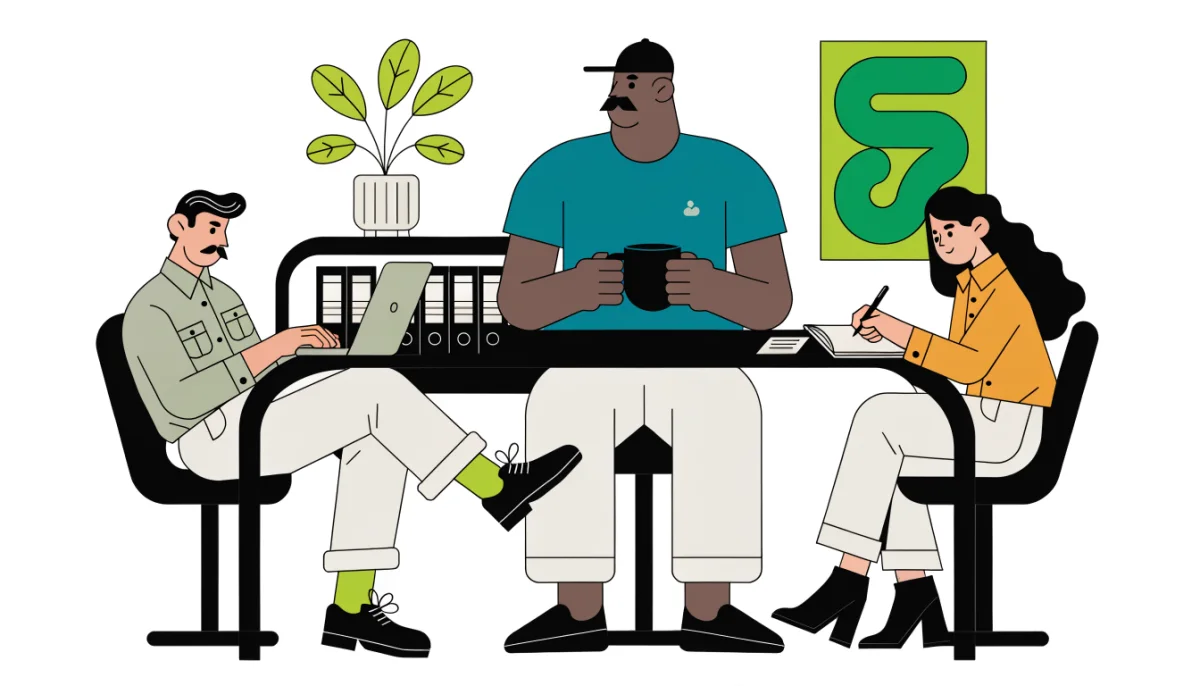 Three colleagues at a desk: one working on a laptop, one standing with a mug, one taking notes